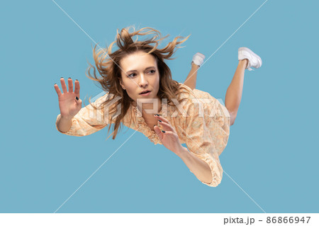 Beautiful young woman levitating in mid-air, falling down and her hair messed up soaring from wind, model flying hovering with dreamy peaceful expression. indoor shot isolated on blue background Beautiful young woman levitating in mid-air, falling down and her hair messed up soaring from wind, model flying hovering with dreamy peaceful expression. indoor shot isolated on blue background 86866947