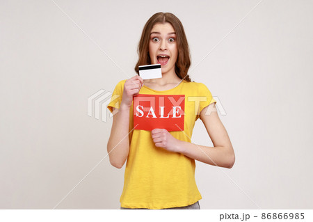 Cheerful excited woman in yellow T-shirt holding showing credit card and card with sale inscription, pay for purchases with credit card, banking. Indoor studio shot isolated on gray background. 86866985