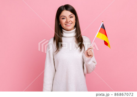 Satisfied positive female holding German flag in her hand and looking at camera with toothy smile, wearing white casual style sweater. Indoor studio shot isolated on pink background. 86867072