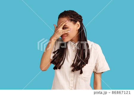 Portrait of interested woman with dreadlocks covering eyes with hand, peeking through fingers with happy curious expression, wearing white shirt. Indoor studio shot isolated on blue background. 86867121