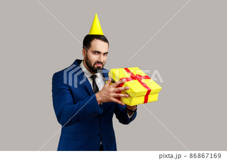Upset man in party cone opening gift box and looking inside with disappointed sad expression, unwrapping bad present, wearing official style suit. Indoor studio shot isolated on gray background. 86867169