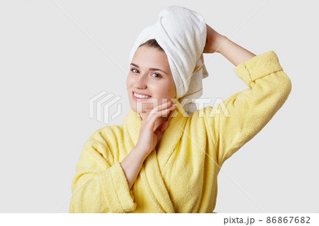 Relaxation and harmony after spa. Young beautiful European woman has towel on head, wears bathrobe, has charming smile, soft healthy skin, models over white studio wall. Natural beauty concept 86867682
