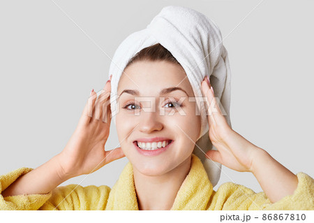 Close up shot of glad satisfied woman being happy after spa procedure, has fresh soft healthy skin, broad smile, white perfect teeth, wears white towel on head, isolated over white background 86867810