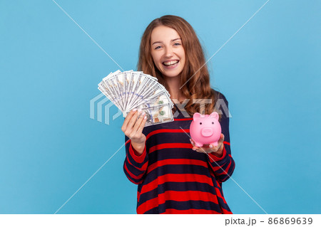Smiling woman wearing striped casual style sweater, holding fan of dollars banknotes and piggy bank, profitable conditions for saving money. Indoor studio shot isolated on blue background. Smiling woman wearing striped casual style sweater, holding fan of dollars banknotes and piggy bank, profitable conditions for saving money. Indoor studio shot isolated on blue background. 86869639