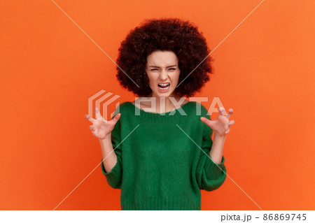 Portrait of angry woman with Afro hairstyle wearing green casual style sweater expressing aggressive emotions, raised arms, screaming. Indoor studio shot isolated on orange background. Portrait of angry woman with Afro hairstyle wearing green casual style sweater expressing aggressive emotions, raised arms, screaming. Indoor studio shot isolated on orange background. 86869745