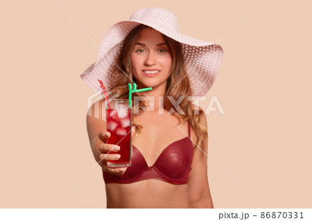 Image of smiling charismatic lady raising one arm, holding glass of red drink with ice, being in good mood, looking directly at camera, being on vacation, wearing light hat and swimming suit. 86870331