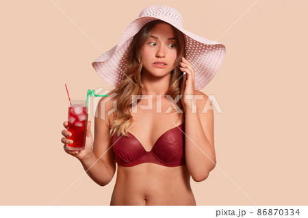 Portrait of surprised attentive lady wearing red swimming suit and hat, standing isolated over beige background, holding red cold cocktail in glass, talking over phone, having important conversation. Portrait of surprised attentive lady wearing red swimming suit and hat, standing isolated over beige background, holding red cold cocktail in glass, talking over phone, having important conversation. 86870334
