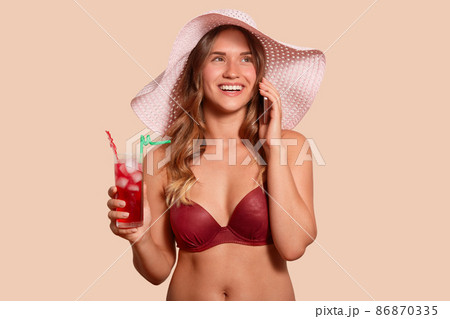 Close up portrait of cheerful magnetic European female being on holidays, wearing swimming suit and hat, holding cocktail and smartphone in both hands, talking over phone. People and emotions concept. 86870335