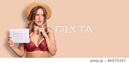 Indoor shot of thoughtful embarrassed female wears summer hat and swimsuit, holds period calendar, tries to remember date of periods, poses against beige background with copy space for your text Indoor shot of thoughtful embarrassed female wears summer hat and swimsuit, holds period calendar, tries to remember date of periods, poses against beige background with copy space for your text 86870397