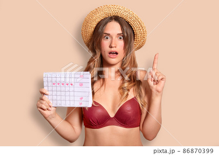 Indoor shot of beautiful European young female has surprised expression, raises index finger, dressed in red bikini and straw hat, holds period calendar, isolated over beige studio background Indoor shot of beautiful European young female has surprised expression, raises index finger, dressed in red bikini and straw hat, holds period calendar, isolated over beige studio background 86870399