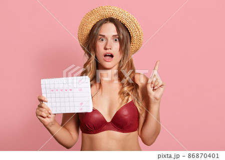 Indoor shot of shocked caucasian young female has surprised facial expression, points up with index finger, wearing in red swimsuit and straw hat, holds period calendar, forget date of periods. 86870401