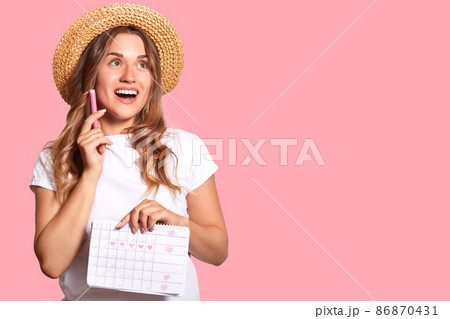 Horizontal shot of positive dreamy female holds marker and periods calendar, being in high spirit, isolated over pink background with copy space for your promotional content or advertisement Horizontal shot of positive dreamy female holds marker and periods calendar, being in high spirit, isolated over pink background with copy space for your promotional content or advertisement 86870431