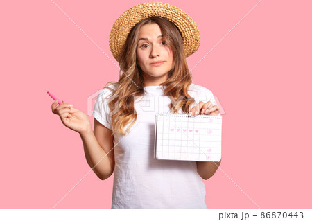 Hesitant young lady carries menstruation period calendar, has clueless expression, wears headgear and white casual t shirt, isolated over pink wall, hesitates about something. Gynecological concept 86870443