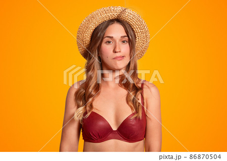 Photo of attractive young female cant stand stuff hot weather conditions during summer, blows air, feels sultry, dressed in swimwear and summer hat, isolated over orange background. Midday sun. 86870504