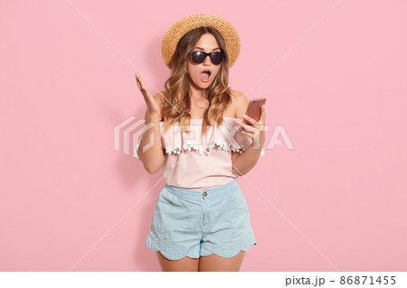 Beautiful astonished woman wearing blouse with bared shoulders, short, straw hat and sunglasses, holding smart phone in hand and posing with open mouth, recives shocking messages from friend. 86871455