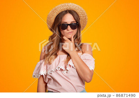 Thoughtful pensive beautiful female holds chin, has hesitant expression, wears shades and summer hat, dressed in shoulder sleeved blouse, poses against orange background. People and rest concept 86871490