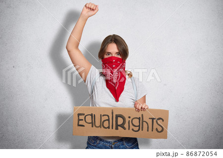 Womens protest concept. Serious young European female with bandana on face, holds plate with inscription, raises hand clenched in fist, isolated over white concrete wall. Feminism and women. 86872054