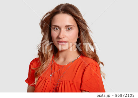 Headshot of gorgeous lovely young female with healthy skin, wavy luxurious hair, dressed in orange sun dress, looks directly at camera, isolated over white background. People and beauty concept Headshot of gorgeous lovely young female with healthy skin, wavy luxurious hair, dressed in orange sun dress, looks directly at camera, isolated over white background. People and beauty concept 86872105