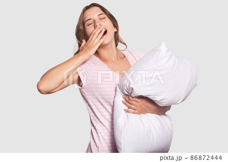 Isolated shot of sleepy woman with widely opened mouth, yawns as hasnt enough sleep, holds soft pillow in hand, isolated over white background. Girl awakes early in morning, Awakening concept 86872444