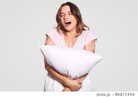 Photo of tired attractive female model yawns, opens mouth widely, dressed in casual clothes, carries pillow, needs to have good rest, isolated over white background. Woman hugs cushion indoor Photo of tired attractive female model yawns, opens mouth widely, dressed in casual clothes, carries pillow, needs to have good rest, isolated over white background. Woman hugs cushion indoor 86872449