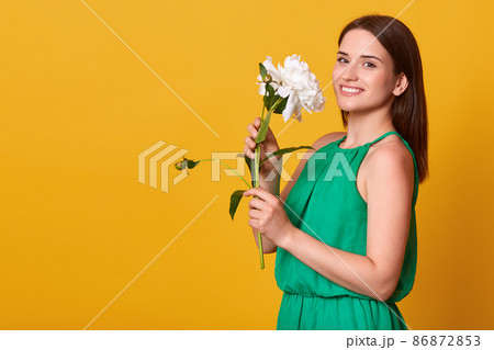 Studio shot of charming woman wearing elegant green sundress isolated over yellow studio background, holding white peony flower, having pleased expression. Copy space for advertisment or promotion. 86872853