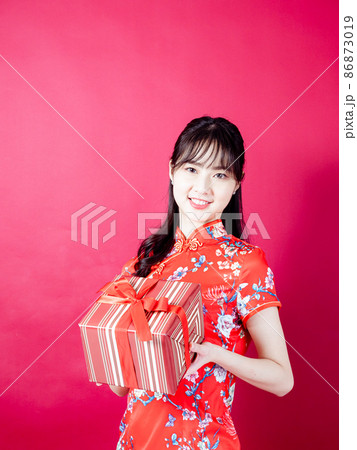 Portrait of a young asian woman wearing traditional cheongsam qipao dress holding gift isolated over red background. 86873019