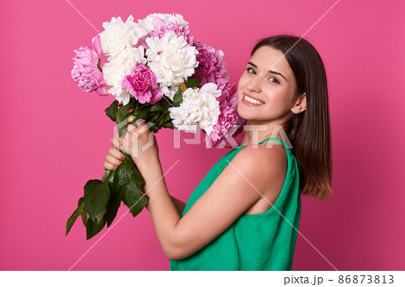 Profile portrait of toothy smiling woman with flowers, dresses green sundress standing over pink background, dark haired female with happy facial expression holds big bouquet of rosy and white peonies 86873813