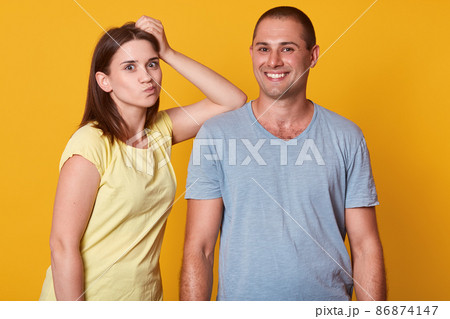 Photo of smiling cheerful man having pleasant facial expression, looking directly at camera. Girlfriend putting her arm on man, looking funny, wearing casual t shirt, isolated over yellow background. 86874147