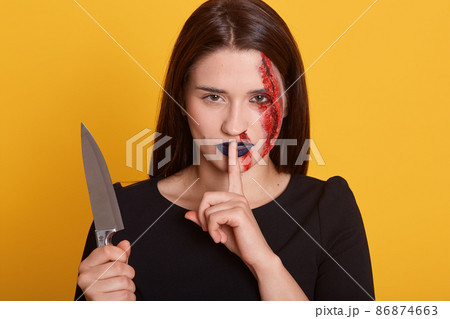 Horizontal shot of young stylish brunette girl with halloween bloody makeup, standing and looking straight with finger near her mouth, showssilent, posing over yellow background. Halloween concept. 86874663
