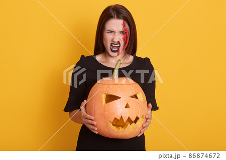 Image of serious young woman wearing black dress, looking screaming directly at camera, lady expresses anger, girl in halloween costume isolated over yellow background with pumpkin in hands. 86874672