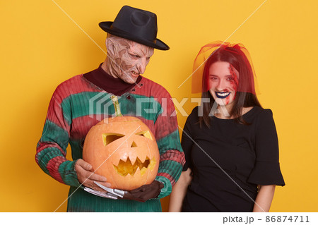 Image of young couple celebrate Halloween, male in striped sweater and black hat, has knives instead of fingers, female in black dress and red veil, isolated over yellow background, guy holds pumpkin. Image of young couple celebrate Halloween, male in striped sweater and black hat, has knives instead of fingers, female in black dress and red veil, isolated over yellow background, guy holds pumpkin. 86874711