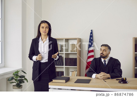 Lawyer or defense attorney making speech during trial session in court. Serious unsmiling woman who represents defendant in lawsuit or criminal prosecution speaking to judge and audience in courtroom Lawyer or defense attorney making speech during trial session in court. Serious unsmiling woman who represents defendant in lawsuit or criminal prosecution speaking to judge and audience in courtroom 86874786
