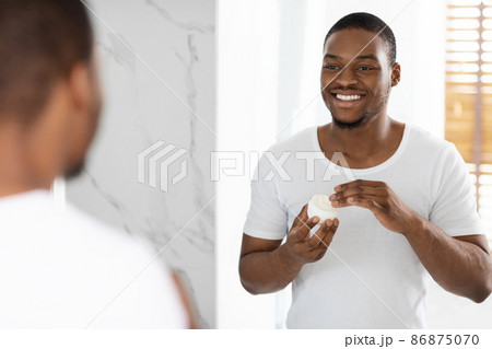 Skincare Concept. Smiling Young Black Man Applying Nourishing Cream On Face 86875070