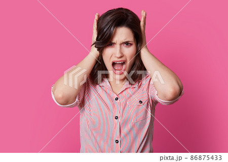 Frustrated brunette lady yells with anger, covers ears, annoyed with loud noise, wears striped shirt, feels outraged, dressed in striped clothes, isolated over bright pink background. Stop this sound Frustrated brunette lady yells with anger, covers ears, annoyed with loud noise, wears striped shirt, feels outraged, dressed in striped clothes, isolated over bright pink background. Stop this sound 86875433