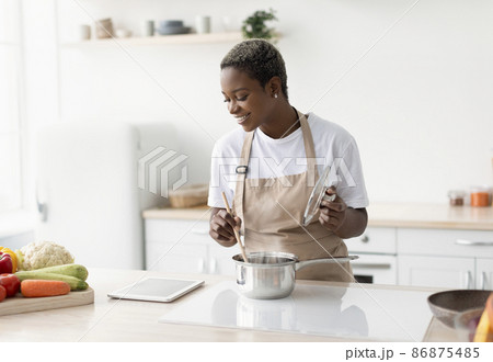 Glad young african american lady in apron cooking and watching video lesson on tablet Glad young african american lady in apron cooking and watching video lesson on tablet 86875485