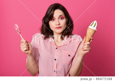 Photo of confused pensive sweet female holding ice cream and lollipop in both hands, looking directly at camera, having bright makeup, making choice, having thoughtful facial expression. Food concept. 86875588