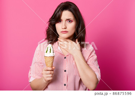 Beautiful girl dressed red and white striped shirt, holds huge ice cream in waffle cone with kiwi taste. Brunette woman keeps hand on neck, lady has sore throat because of eating lots cold sundae. 86875596