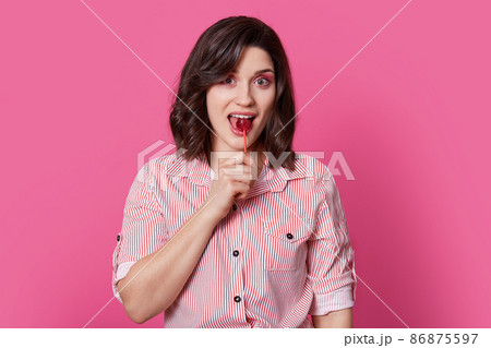 Portrait of happy pleased brunette woman with wavy hair, has pink makeup, licks lollipop, wears striped fashionable shirt, isolated over bright rosy background, being sweet tooth. Pinup girl indoor 86875597