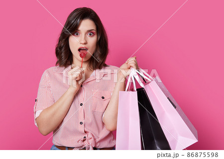 Isolated shot of pinup girl with wavy hair, has surprised expression, enjoys sweet lollipop, holds bags, makes shopping, dressed in stylish shirt, isolated over pink background. Coquettish woman 86875598