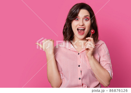 Horizontal shot of joyful attractive woman with makeup, holds lollipop, keeps mouth widely opened, wears stylish outfit, points with thumb aside, shows blank space for your advertisement or text Horizontal shot of joyful attractive woman with makeup, holds lollipop, keeps mouth widely opened, wears stylish outfit, points with thumb aside, shows blank space for your advertisement or text 86876126