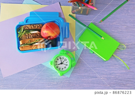 Lunchbox with cereal bread sandwich with cheese, organic vegetables and micro greens, apple, stationery and alarm clock on wooden table, top view 86876223