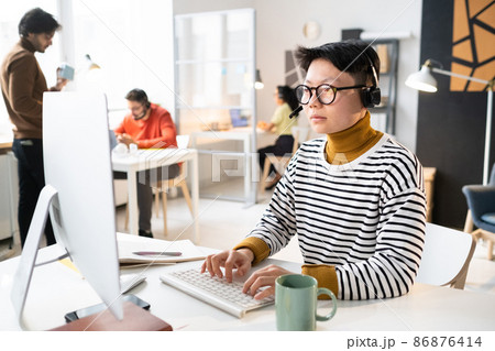 Asian operator in headphones sitting at the table with computer and typing on keyboard, she working with clients online 86876414