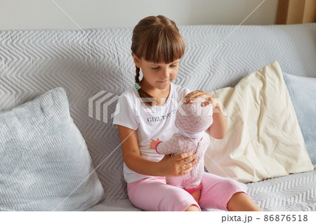 Photo of preschooler girl sitting on the sofa in the room at home playing with baby doll like mom, child wearing white t shirt and pink short, childhood. Photo of preschooler girl sitting on the sofa in the room at home playing with baby doll like mom, child wearing white t shirt and pink short, childhood. 86876518