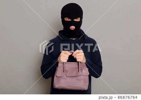 A young guy thief is dressed in a black hoodie and robbery mask, holds a woman's bag with both hands, stole other people's things, looks at the camera, isolated on gray background. 86876764
