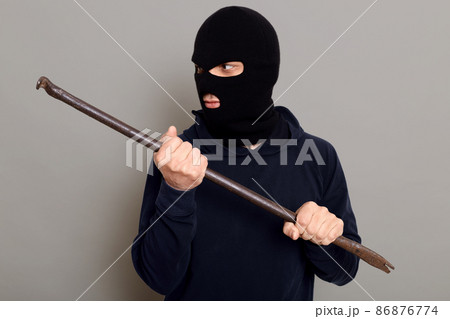 Man thief with handy crowbar looks away, wants to break open door or lock, dresses in balaclava and turtleneck, dangerous bandit commits a crime, posing isolated over gray background. 86876774