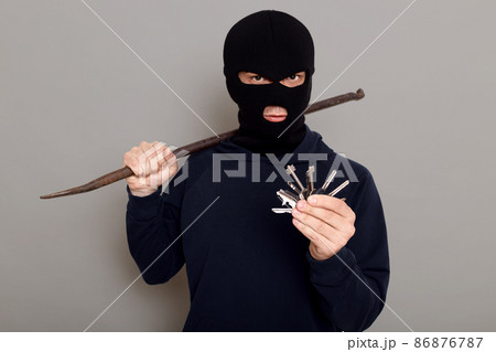 Criminal thief holds iron crowbar and bunch of keys in his hands, being about to rob apartment, looks at camera with insidious expression on his face, isolated over gray background. 86876787
