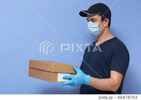 Profile of handsome careful delivery man holding parcel, having working day, wearing uniform and antibacterial mask, gloves, standing isolated over blue background in studio. Quarantine concept. Profile of handsome careful delivery man holding parcel, having working day, wearing uniform and antibacterial mask, gloves, standing isolated over blue background in studio. Quarantine concept. 86878703