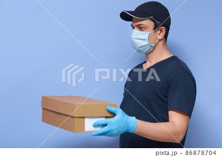 Indoor shot of young delivery man with carton box in hands, male wearing t shirt, cap and protective mask and latex gloves, meet his client to give order,posing isolated over blue studio background. Indoor shot of young delivery man with carton box in hands, male wearing t shirt, cap and protective mask and latex gloves, meet his client to give order,posing isolated over blue studio background. 86878704