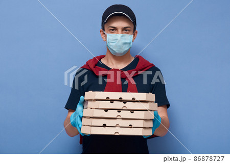 Horizontal indoor picture of charismatic pleasant delivery guy holding ordered food in boxes, wearing gloves and antibacterial mask, having peaceful facial expression. Quarantine time concept. 86878727