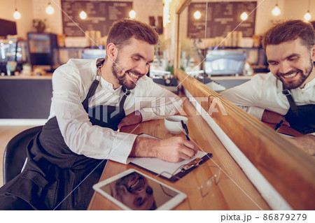 Cheerful male worker writing on clipboard in coffee shop Cheerful male worker writing on clipboard in coffee shop 86879279